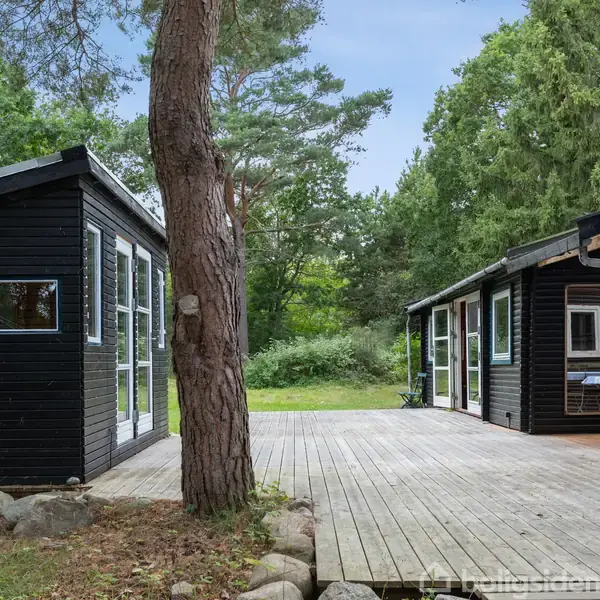 Sort træsommerhus står stille ved siden af et stort, åbent terrasseområde belagt med træbrædder, omkranset af frodig, grøn skovvegetation under en klar blå himmel.