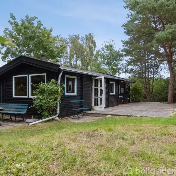 Sort træhus med hvide vinduer og dør står stille omringet af træer og græs. Et havebord med bænke står på fliseterrassen foran huset.