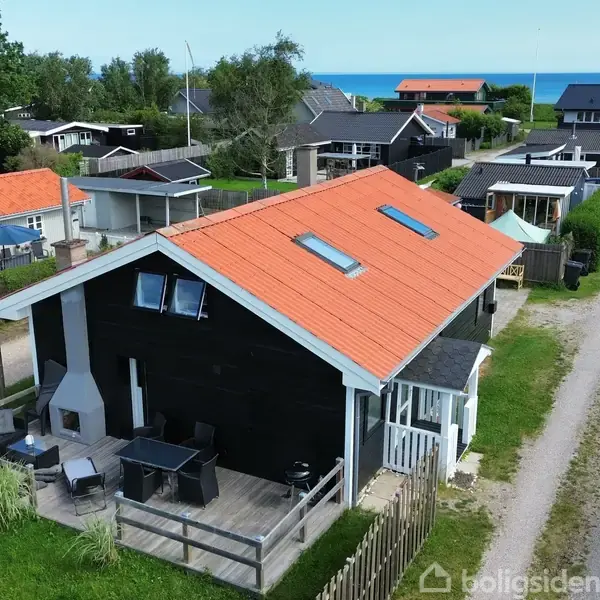 Et sort træhus med orange tag og ovenlysvinduer står stille. Det ligger i et sommerhusområde ved en grusvej, omgivet af hegn og tæt på havet.