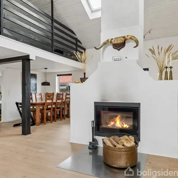 Pejsen brænder i et lyst, moderne stueområde med et spisebord i baggrunden. Der er en overetage med sort gelænder, og rummet er indrettet med planter og træmøbler.
