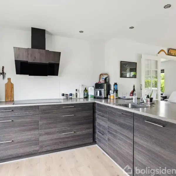 Køkken med mørke træskabe og gråt bordplade; stålvask og emhætte. Knive hænger på væggen, og der er diverse køkkenredskaber og flasker. Lys træparketgulv og hvide vægge.