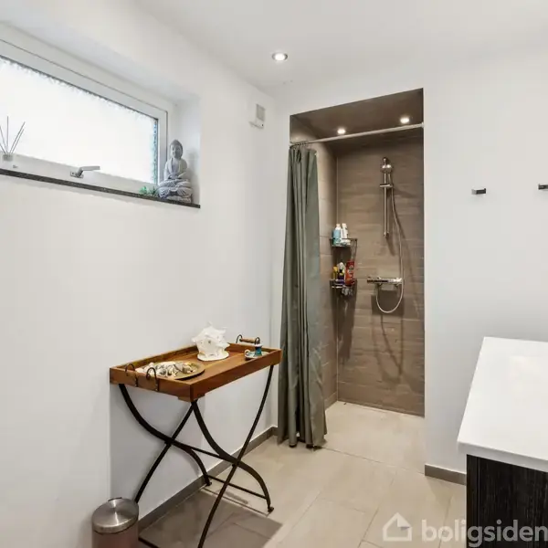 Et moderne badeværelse med brusekabine til højre, indrettet med lyse fliser. Et bord til venstre holder genstande, mens et vandret vindue lukker lys ind.