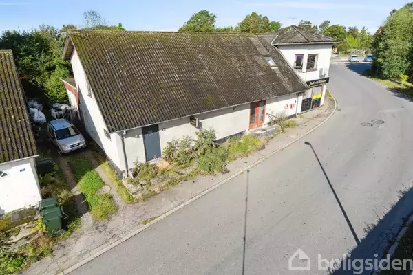 Bygning ved en vejkurve, lukket dør og planter foran. Hus med skråt, ældre tag og lidt vegetation langs siden. Bilen er parkeret ved siden af huset.