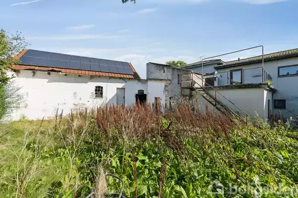 Hus med solpaneler på taget står i en have med højt græs og vilde planter. En udendørs trappe fører til en balkon på husets side.