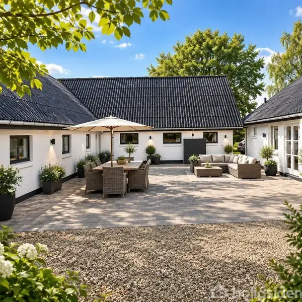 En gårdhave med stenbelægning har et bord og stole under en parasol samt en sofa med sofabord. Omkranset af hvide bygninger med sorte tage og grønne planter.