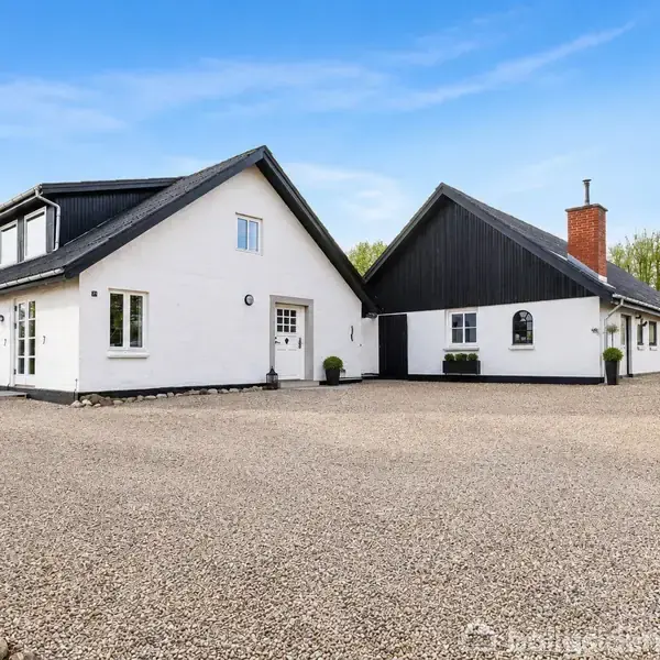 Hvidt hus med sort tag og flere vinduer står på en grusbelagt gårdsplads, omgivet af træer i baggrunden under en blå himmel.