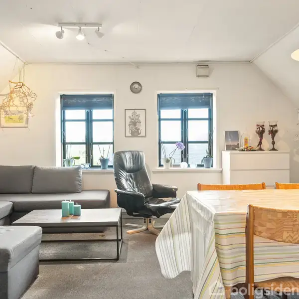 Stue med grå sofa og lænestol ved siden af et sofabord, beliggende nær to vinduer. En spisebord med dug og stole er i rummet, hvor der også findes dekorative elementer.