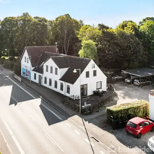 En hvid to-etagers bygning står langs en vej i et skovområde. Foran bygningen er en parkeringsplads med en rød bil. Baggrunden er præget af mange træer.