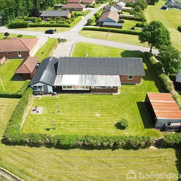 Hus set fra oven med sort tag og tilstødende garage. Omkranset af klippet græsplæne og hæk. Omgivelserne inkluderer nabohuse og veje i en villakvarter.