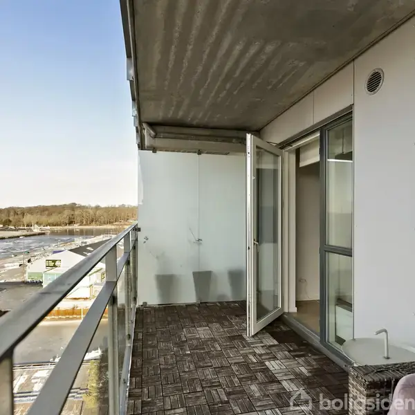 En altan med trægulv, hvor døren står åben til en lejlighed. Altanen har et glasgelænder og udsigt over et industriområde samt træer i horisonten.