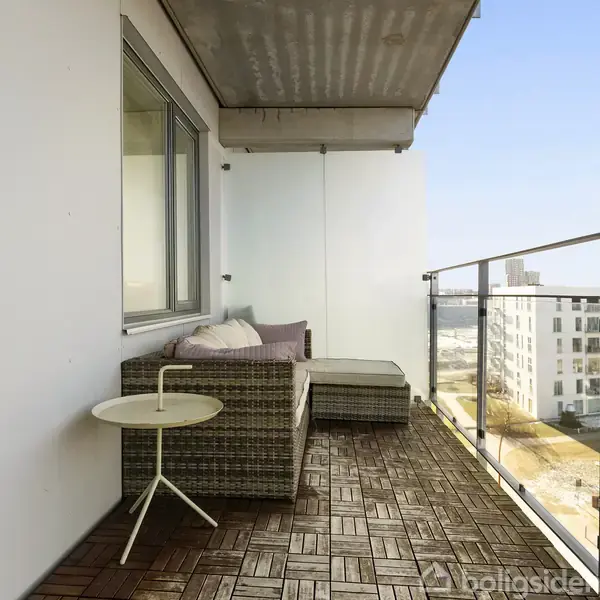 En balkon med træfliser har en fletsofa med puder og et lille rundt bord. I baggrunden ses moderne bygninger og en blå himmel.