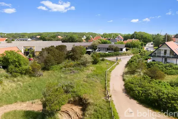 En snoet landevej løber gennem en grøn, landlig bebyggelse omkranset af buske og små træer, med huse i baggrunden under en klar blå himmel.