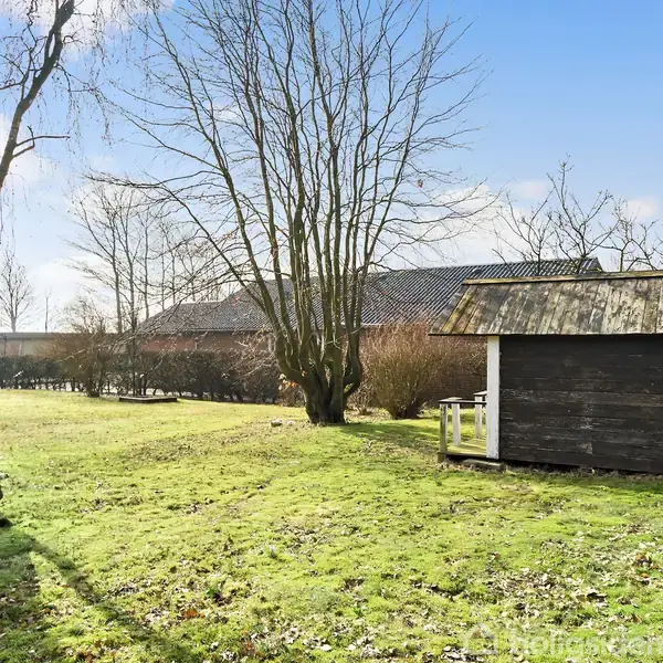 Et lille træhus står stille på en græsplæne omgivet af træer og en hæk. I baggrunden ses en murstensbygning under en klar blå himmel.