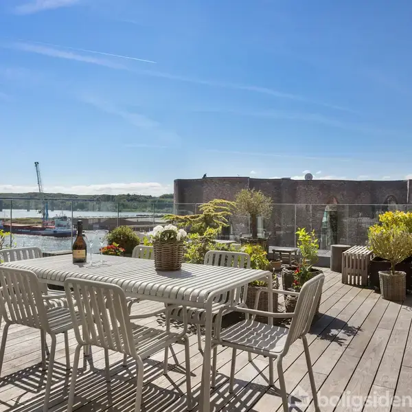 En udendørs terrasse med et bord og stole står omgivet af blomster og planter i krukker. I baggrunden ses en industriel bygning ved vandet under en klar blå himmel.