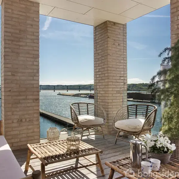 En altan med havemøbler og planter vender ud mod en marina med både og en bro i baggrunden. Sollyset belyser området, og der er udsigt over vandet.