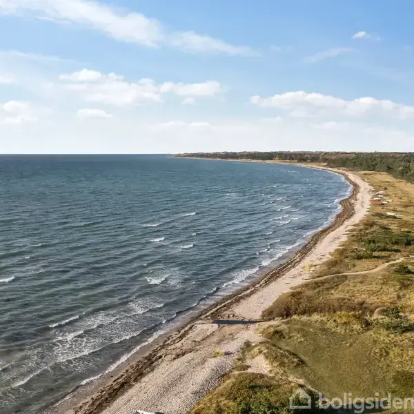 Kystlinje langs en bølget havoverflade strækker sig mod horisonten. Sandet strand med grus er omgivet af græs og træbevoksning under en blå himmel med lette skyer.