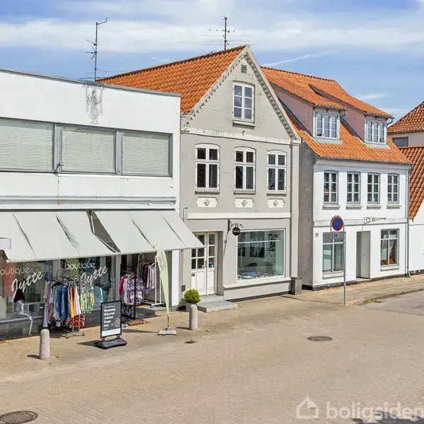 Et række af butikker langs en byvej, hvor en butik udstiller tøj udenfor. Forretningerne har traditionelle bygninger med orange tegltage og lys facade. En bil ses parkeret til højre. Senbrug, boutique Julyte.