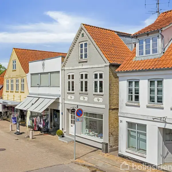 Et rækkehus med rødt tegltag står i en rolig bygade, omgivet af andre huse med butikker i stueetagen. En vejskiltning forbyder parkering foran bygningen.