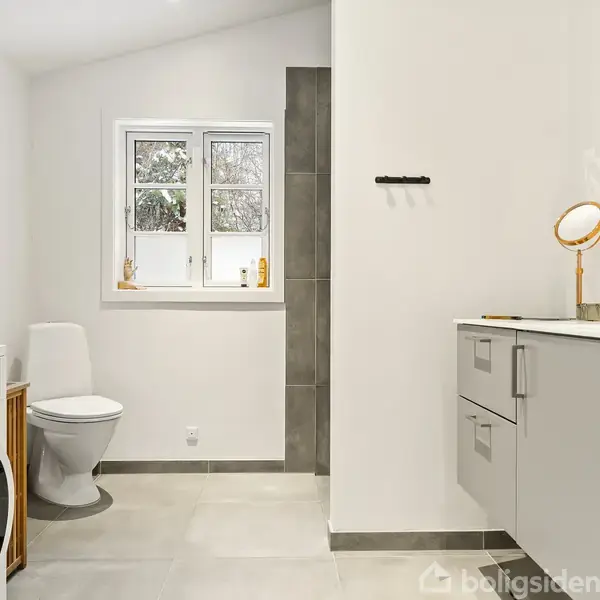 Et moderne badeværelse med grå skabe og spejl. Til venstre står et toilet ved siden af en vaskemaskine. Et vindue med udsigt til sneen giver lys.