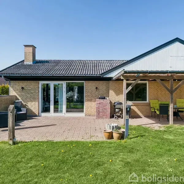 Et en-etagers hus med gul murstensfacade står i en have. En terrasse forbundet til huset har borde, stole og en parasol under en overdækket sektion.