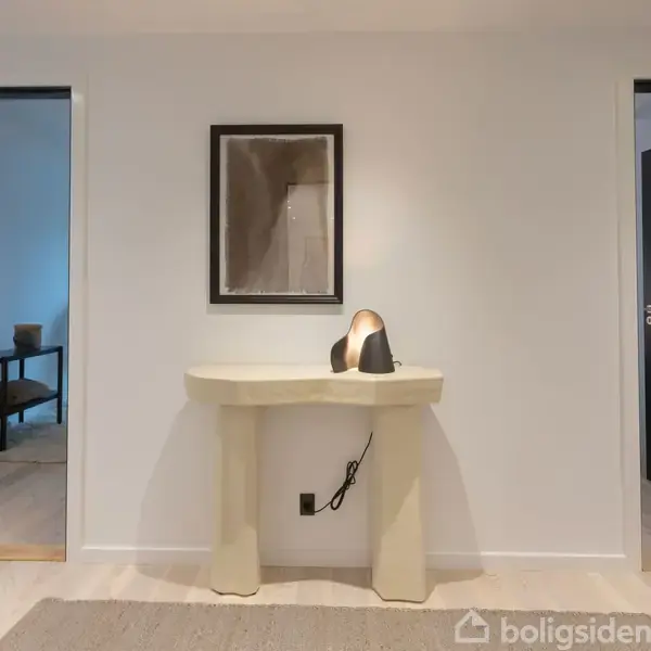 Et beige konsolbord står i midten af en hvid væg med en bordlampe placeret ovenpå. På væggen hænger et indrammet billede. To døråbninger viser tilstødende værelser.