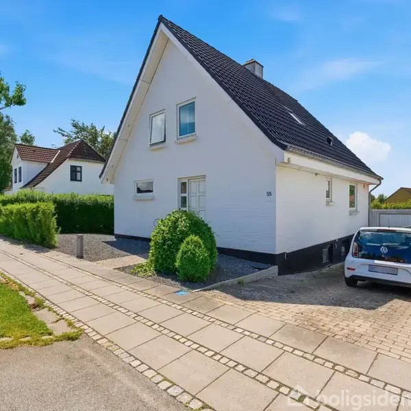 Et hvidt hus med sort tag har en bil parkeret i indkørslen. Det står i et villakvarter med grønne hække. Skilt fra "Rell Ejendomsmægler - Til salg".