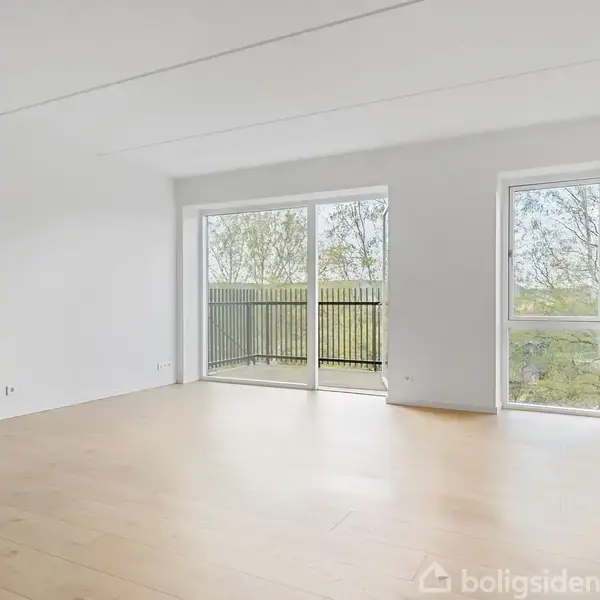 Et tomt rum med lyse trægulve og hvide vægge. En glasdør åbner til en balkon med udsigt til træer udenfor.