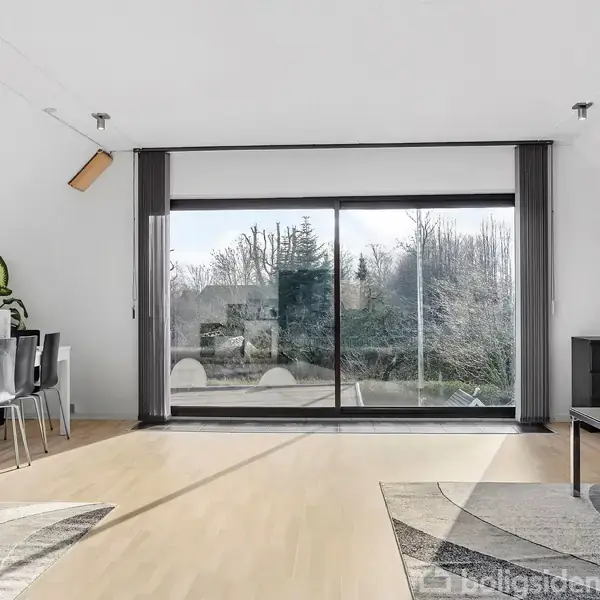 En lys stue med et spisebord til seks personer til venstre. Store glasdøre åbner ud mod en terrasse med udsigt til træer. Rummet er enkelt og moderne indrettet.