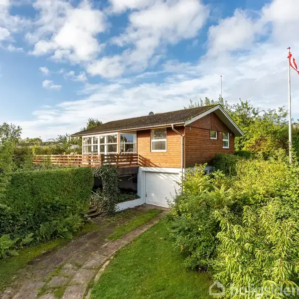 Et træhus står på en grøn grund med en flagstang til højre. Huset har en veranda, omgivet af buske og træer under en blå himmel med skyer.