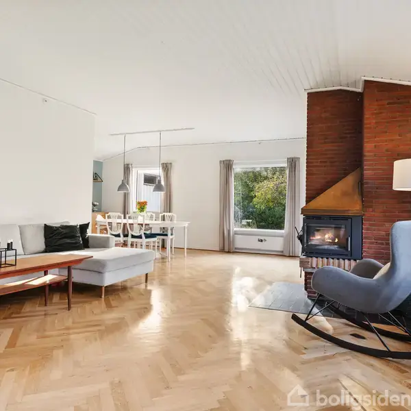 Grå hjørnesofa står i lys stue med trægulv, ved siden af en åben pejs med rød murstensramme. Spisebord og store vinduer giver udsigt til grønne omgivelser.