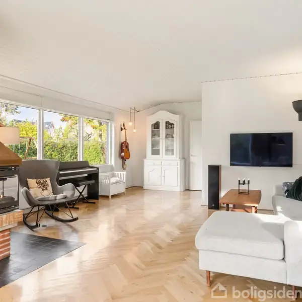 En lys stue med en hvid sofa og et fladskærms-TV, omgivet af store vinduer. En pejs er integreret i en murstensvæg. Et klaver og guitar står nær hjørnet.