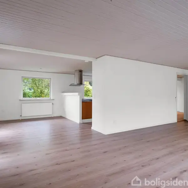 Stue med trægulv og hvide vægge. Køkken ses gennem åbent område, mens et vindue giver udsigt til træer udenfor. Døren fører til et tilstødende rum.