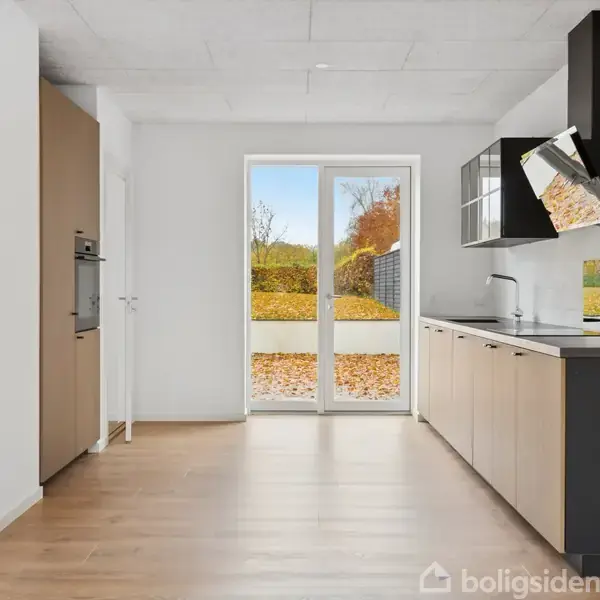 Køkken med moderne skabe og indbygget komfur ved siden af en glasdør. Udenfor ses efterårsblade på jorden og grønne buske. Pæne lyse vægge og trægulv.