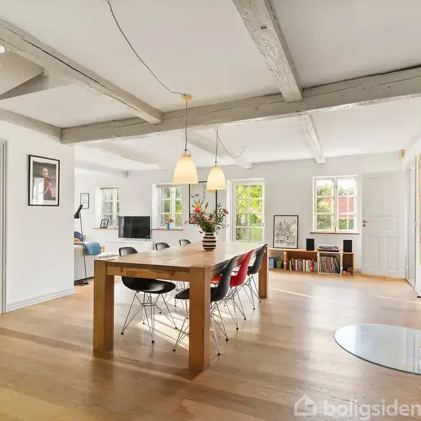 Spisebord med stole står i en lys stue med trægulv. Brændeovn til højre, store vinduer, loftbjælker, og lamper skaber en hyggelig atmosfære. I baggrunden ses flere møbler.