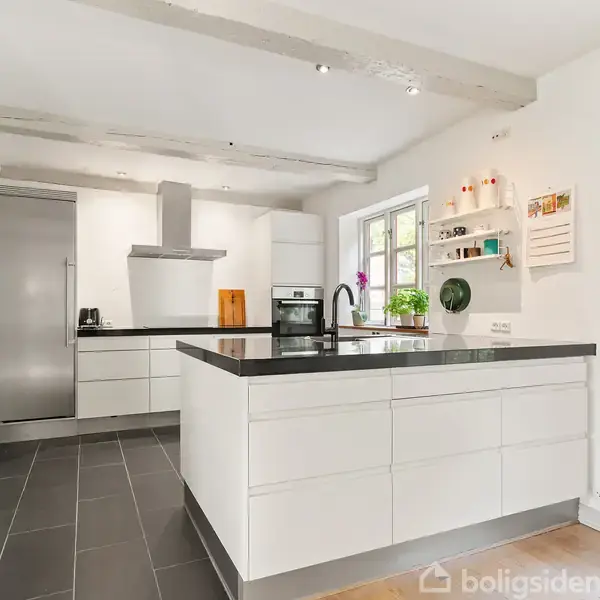 Et moderne køkken med en stor, hvid køkkenø og sort bordplade. Der ses en indbygget ovn og et højskab til venstre. Rummet er lyst med hylder og vindue.