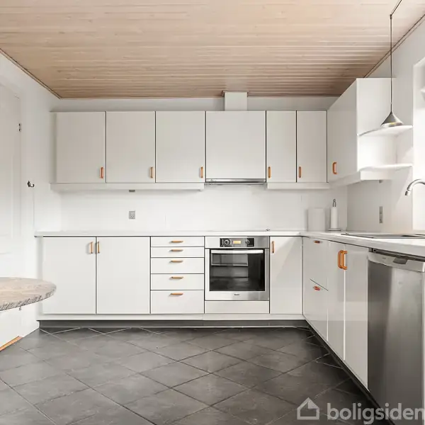 Et moderne køkken med hvide skabe og skuffer, sort kakkelgulv og træloft. En indbygget ovn og opvaskemaskine er synlige. Et rundt bord med sorte ben står til venstre.