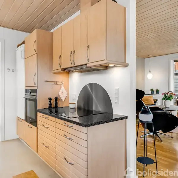 Køkken med træskabe og sort bordplade, ovn og emhætte; døråbning fører til stue med træmøbler og store vinduer; lys og moderne indretning.