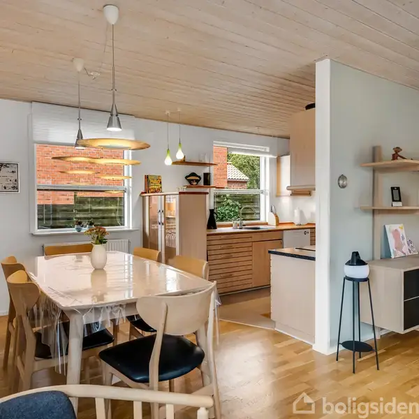 Spisebord med stole står midt i et lyst køkken-alrum. Bordet er dækket med voksdug, og en vase med blomster pynter. I baggrunden ses et køkken med træpaneler og skabe.