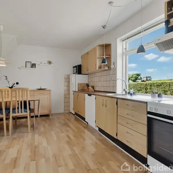 Køkken med ovn og træskabe langs væggen. Spisebord til venstre under en lampe. Store vinduer med udsigt til en grøn hæk udenfor. Klædt i lyse træfarver.