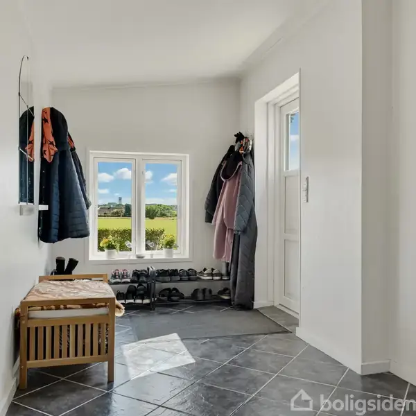 En entré med flisegulv har en bænk og tøj hængende på kroge. Der er sko arrangeret ved et vindue med udsigt til en grøn mark og blå himmel.