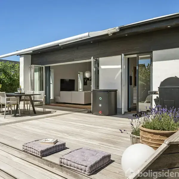 Terrasse med træmøbler står stille i solen foran et moderne, åbent hus. Planter i krukker omkranser området, og dørene til stuen er åbne, afslørende et stilfuldt interiør.