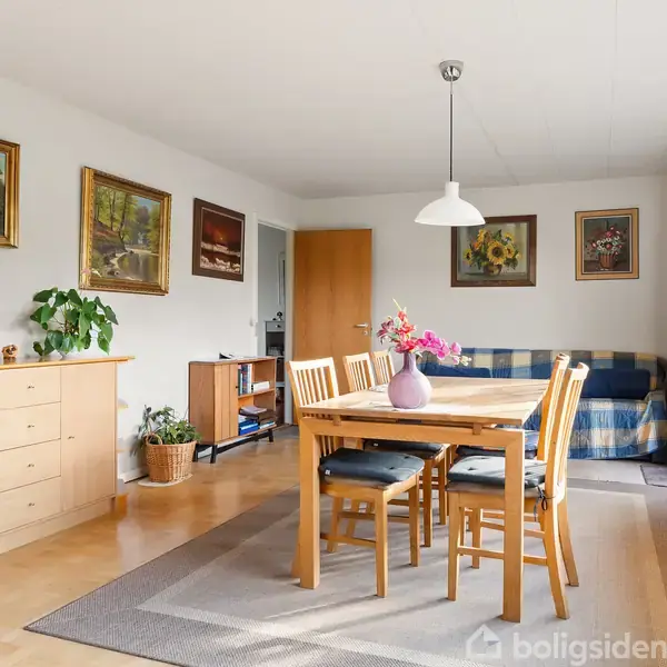 Spisebord med træstole står på et tæppe i en lys stue. Væggene har malerier, og en sofa er i baggrunden. Store vinduer giver naturligt lys.