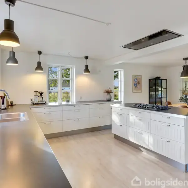 Køkken med en kogeø i centrum, omgivet af moderne skabe og vinduer. Lys hænger fra loftet, og der er en spiseplads i baggrunden med stole og bord.