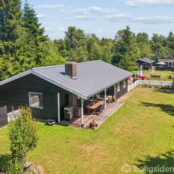 Et sort træhus med en overdækket terrasse står i en græsbelagt have omgivet af træer. Huset har en skorsten, og der ses flere mindre bygninger i baggrunden.