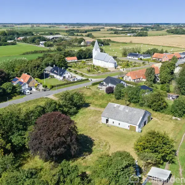 En hvid landbygning står stille omgivet af grønne marker og træer. I baggrunden kan man se en lille landsby med en karakteristisk hvid kirke.