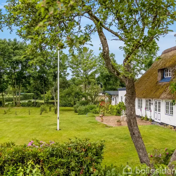 Hvidt hus med stråtag står stille omgivet af en frodig have med træer og blomstrende buske. Flagstang ses i forgrunden, og himlen er blå med spredte skyer.