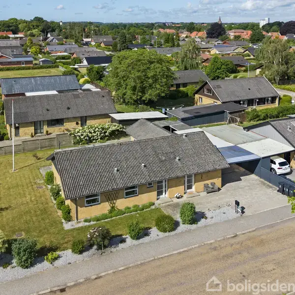 Flere enfamiliehuse med flade tage og gule mursten ligger i en villavej. Nogle haver har buske og stier. Biler holder parkeret under carporte; vejen er løbet med træer og grønne områder.
