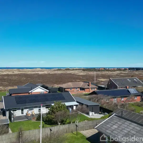 Huse med solpaneler står stille blandt græsplæner og hegn, med en åben udsigt over en hede der strækker sig mod en fjern horisont ved havet under en blå himmel.