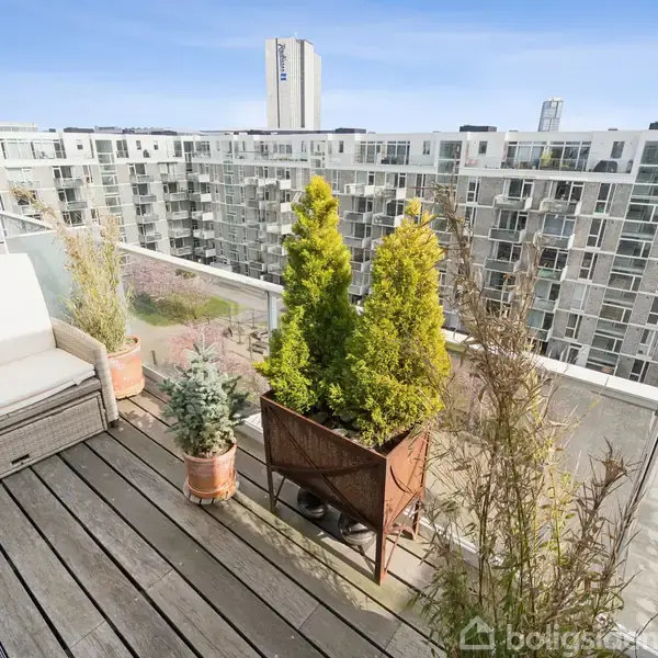 En altan med trædækning har forskellige grønne planter i krukker ved siden af en sofabænk. Udsigten viser flere etagebygninger og en klar blå himmel i et bymiljø.