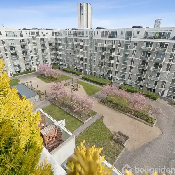 En stor, moderne boligblok omkranser en indre gårdhave med blomstrende træer og stier. Udsigt fra en højere etage, tydelig blå himmel og høje bygninger i baggrunden.